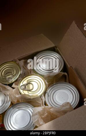 Una scatola di cartone aperta piena di lattine di stagno. I raggi del mattino illuminano una varietà di cibo in scatola in lattine piene di stagno. Cibo in scatola. Vista dall'alto ad angolo. SEL Foto Stock