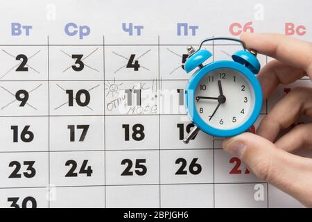 Una mano trattiene una piccola sveglia, in background è un calendario con il giorno segnato e la parola vacanza Foto Stock