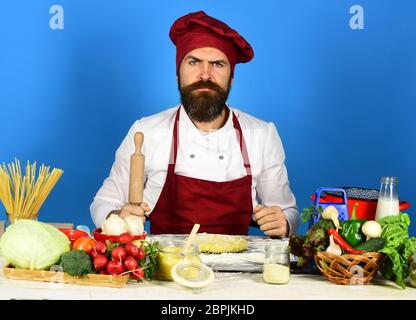 Cuocete con il viso serio in uniforme borgogna tiene il perno di rotolamento. L'uomo con la barba si siede sul piano di appoggio su sfondo blu. Chef con pasta, verdure e pasta sul tavolo. Concetto di cibo fatto in casa. Foto Stock