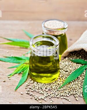 Olio di canapa in due vasi di vetro con grano nel sacco e sul tavolo, foglie di cannabis sullo sfondo di un vecchio pannello di legno Foto Stock