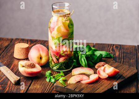 Bottiglia di acqua infusa con pesche tagliate a fette e le foglie di basilico. Coltello e ingredienti sul tagliere. Foto Stock