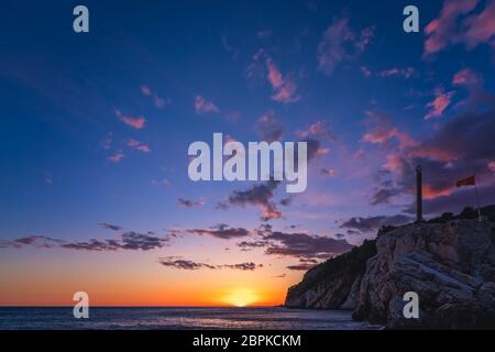 Statua obelisco e bandiera montenegrina che fluttua nel vento nella fortezza sulla cima della scogliera a Petrovac costa al tramonto, Montenegro Foto Stock