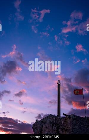 Statua obelisco e bandiera montenegrina che fluttua nel vento nella fortezza sulla cima della scogliera a Petrovac costa al tramonto, Montenegro Foto Stock