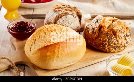 Tutta la panini di vari tipi di pane fresco servito nel ristorante sul tagliere di legno, con burro e marmellata in ciotole in vetro Foto Stock