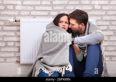 Coppia giovane avvolto in una coperta seduti nella parte anteriore del radiatore contro un muro di mattoni a casa Foto Stock