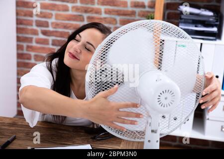 Sorridente giovane donna godendo brezza con ventola elettrica in ufficio Foto Stock