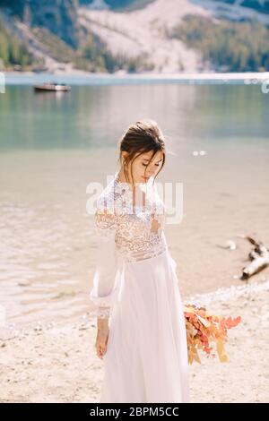 Bella sposa in abito bianco con maniche e pizzo, con bouquet giallo autunnale di fiori secchi e rose di pony, sul Lago di Braies in Italia Foto Stock