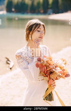 Bella sposa in abito bianco con maniche e pizzo, con bouquet giallo autunnale di fiori secchi e rose di pony, sul Lago di Braies in Italia Foto Stock