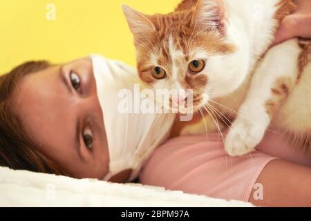 Giovane donna in bianco maschera virus viso di stoffa giocando con il suo gatto, dettaglio su occhi felini, proprietario sfocato in background Foto Stock