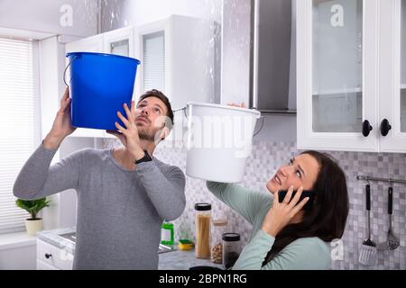 Preoccupato per il giovane donna chiamando l'idraulico sul cellulare mentre l'uomo è la raccolta di acqua di perdita dal soffitto Foto Stock