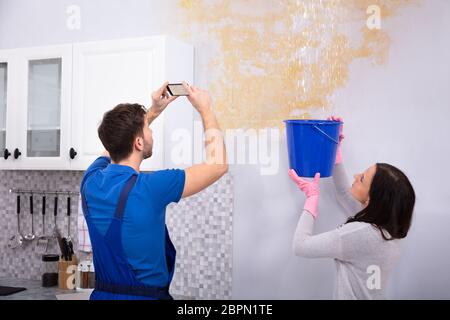 Donna raccolta di acqua in blu la benna da soffitto danneggiato mentre Repairman tenendo la fotografia sul Mobilephone Foto Stock