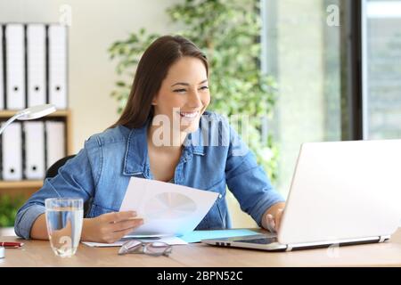 Imprenditore tenendo una lavorazione carta confrontando i documenti in linea con un computer portatile in ufficio Foto Stock