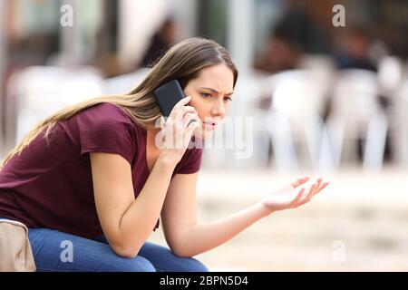 Arrabbiato casual donna chiamando al telefono seduta su una panchina in strada Foto Stock