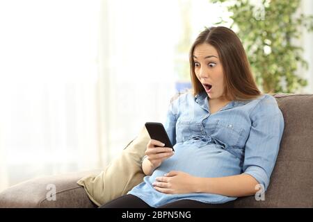 Stupiti donna incinta la visione di contenuti multimediali in uno smart phone seduto su un divano nel soggiorno in un interno di una casa Foto Stock