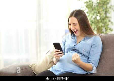 Excited donna incinta la visione di contenuti multimediali in linea in un telefono cellulare seduta su un divano nel salotto di casa Foto Stock