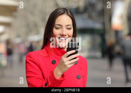 Donna felice indossando giacca rossa passeggiate utilizzando smart phone in inverno in strada Foto Stock