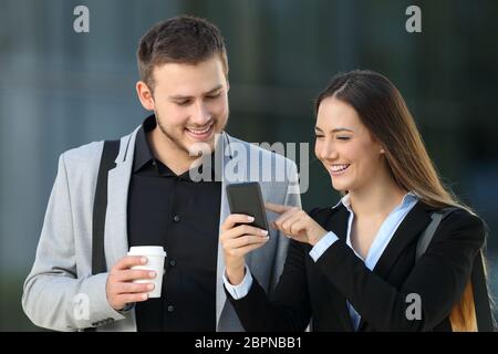 Vista frontale di due dirigenti felici che parlano di contenuti per smartphone in piedi all'aperto per strada Foto Stock