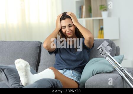 Vista anteriore verticale di un triste donna disabile con piede di gesso lamenta seduta su un divano nel salotto di casa Foto Stock