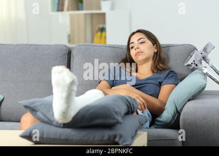 Donna disabile con un piede in gesso noioso seduta su un divano nel salotto di casa Foto Stock