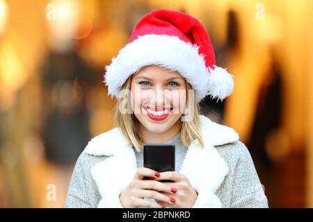 Ritratto di una bella donna che indossa il cappello di babbo natale durante le vacanze di natale con smartphone sulla strada Foto Stock