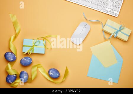 Uova di pasqua colorate, confezione regalo, tastiera e mouse per computer, foglio vuoto con busta su sfondo giallo. Vista dall'alto con spazio per la copia. Biglietto di auguri Foto Stock