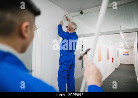 Elettricista Senior sta parlando al suo partecipante durante il montaggio di una telecamera di sicurezza nel corridoio di un edificio per uffici. Foto Stock