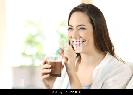 Donna felice di bere cacao agitare guardando la telecamera seduta su un divano nel salotto di casa Foto Stock