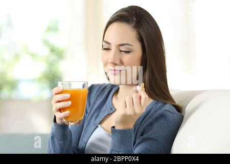 Donna che mantiene un succo di arancio e vitamina pillola seduta su un divano nel salotto di casa Foto Stock