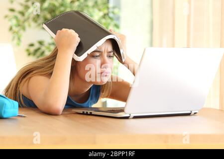 Annoiato studente facendo i compiti on line con un laptop soffiando con un notebook sulla testa in una tabella a casa Foto Stock