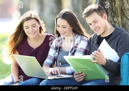 Tre felici gli studenti che studiano in linea con un computer portatile e leggere note di seduta sul prato in un parco Foto Stock