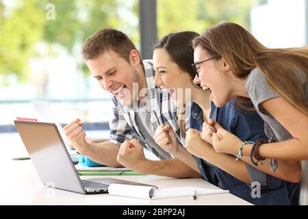 Vista laterale di tre studenti entusiasti la lettura di buone notizie insieme sulla linea in un laptop seduto in una scrivania in una classe Foto Stock