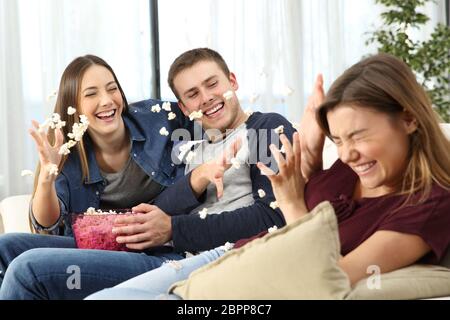 Tre amici felici scherzando e gettando popcorn seduto su un divano nel soggiorno di casa Foto Stock