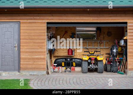 Facciata vista frontale porta aperta ATV quad moto parcheggio garage disordinato edificio con lato in legno a casa strada cortile e prato percorso. Casa Foto Stock