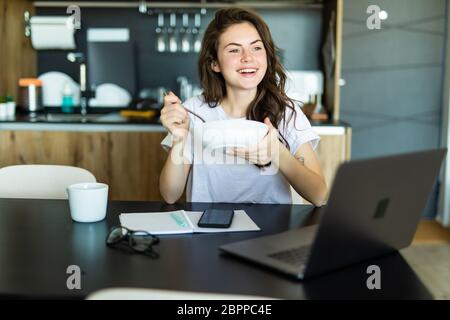 Una donna attraente che mangia la colazione e lavora o che si occupa dei social media sul suo computer portatile si siede al tavolo da pranzo Foto Stock