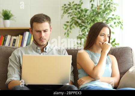 Marito che usa un computer portatile in linea ignorando la sua triste moglie seduta su un divano a casa Foto Stock