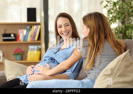 Felice donna incinta a parlare con un amico che tocca il suo ventre seduta su un divano nel salotto di casa Foto Stock