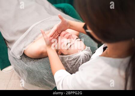 Il medico estetista effettua un massaggio cosmetico del viso. Donna si rilassa su una sedia cosmetica Foto Stock