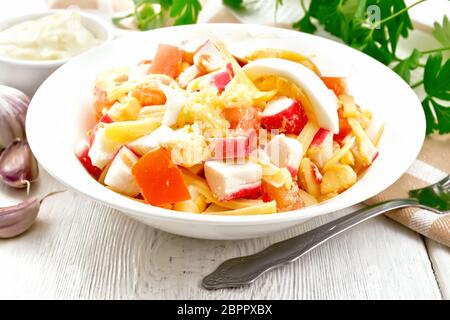 Insalata di bastoncini di granchio, formaggio, aglio, pomodori e uova con la maionese in una piastra, asciugamano, il prezzemolo e la forcella sullo sfondo della luce di tavole di legno Foto Stock