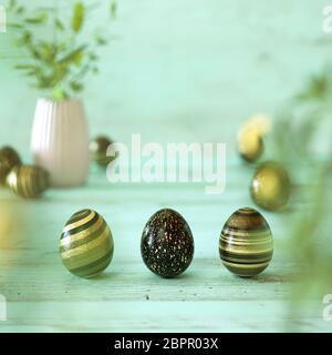 Tre di oro nero e uova di Pasqua decorate con strisce, splatter, fiori e macchie in oro nero e giallo su verde menta sullo sfondo di legno con copia Foto Stock