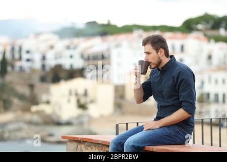 Vista laterale verticale di un uomo rilassato che guarda lontano di bere il caffè seduti su una mensola in vacanza in una città costiera Foto Stock