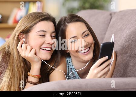 Vista frontale ritratto di due amici gioioso ascolto di musica on line da uno smart phone giacente su un divano nel salotto di casa Foto Stock