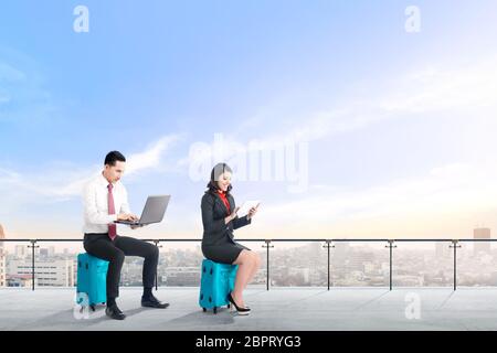 Due giovani asian business gente seduta sulla valigia blu tenere il laptop e tablet lavorando sulle moderne di terrazza con vista sulla città e cielo blu sullo sfondo Foto Stock