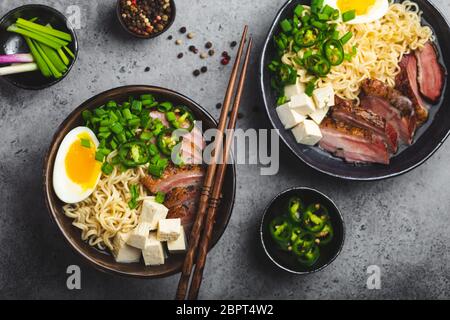 Due ciotole di gustosa zuppa di noodle asiatica ramen con brodo di carne, tofu, maiale, uovo con tuorlo su fondo grigio rustico in cemento, primo piano, vista dall'alto. Colata calda Foto Stock