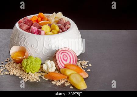 Pasto di organico per animali domestici mescolati in bianco ciotola per gatto o cane, costituito da carne, uova crude e verdure fresche, visto da vicino contro backgroun nero Foto Stock