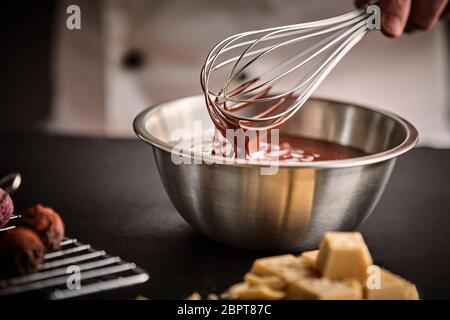 Lo Chef tenendo una frusta con il cioccolato fuso in un recipiente di miscelazione in cucina con ingredienti e bomboni a fianco Foto Stock