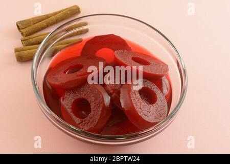 Anelli di mele speziati rossi in una ciotola di vetro con bastoncini di cannella su sfondo rosa Foto Stock