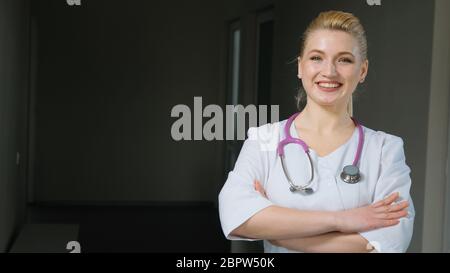 Bionda in camice medico bianco e stetoscopio viola ruota la testa verso la fotocamera, piega le braccia insieme. Dipendente femminile positivo con volto sorridente Foto Stock