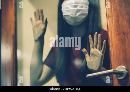 Closeup mano di donna asiatica che indossa maschera medica che tocca porta di vetro quando rimanere isolamento a casa per auto quarantena in Covid-19 focolaio situazione, c. Foto Stock