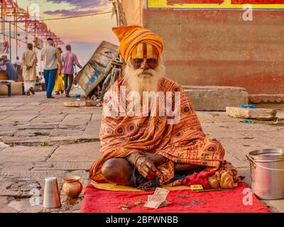 Varanasi, India - 13 novembre 2015. Un sadhu anziano si siede su un tappetino vicino ai Ghat del fiume Gange, indossando abiti tradizionali e contrassegni indù della fronte. Foto Stock