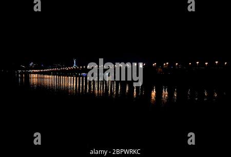 Vista notturna del ponte Paton attraverso l'ampio fiume Dnieper e le luci della riva destra di Kiev. Le lanterne di un lungo ponte si riflettono in Foto Stock